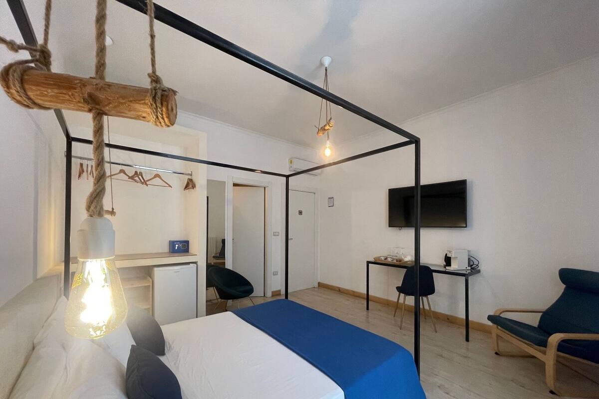 A modern hotel room with a bed, a desk, a chair, a TV, and a hanging light fixture.