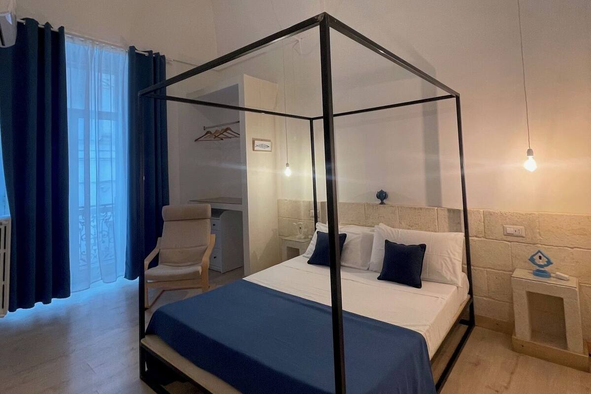 A bedroom with a four-poster bed, a bedside table, a chair, and a window with curtains.