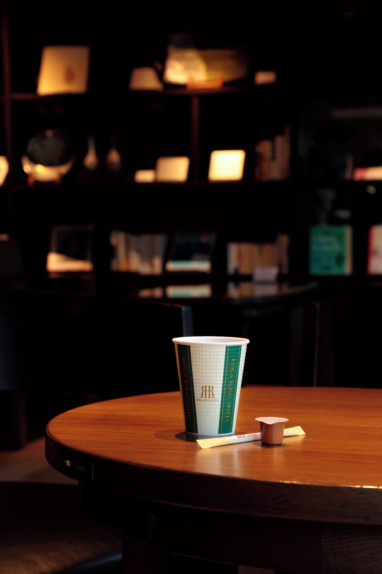 Una mesa con una taza de papel etiquetada "Riga Royal Hotel" y un pequeño recipiente, posiblemente para salsas o servilletas, sobre una mesa de madera en una habitación poco iluminada con varias mesas y sillas, lo que sugiere un área de comedor o de reuniones.