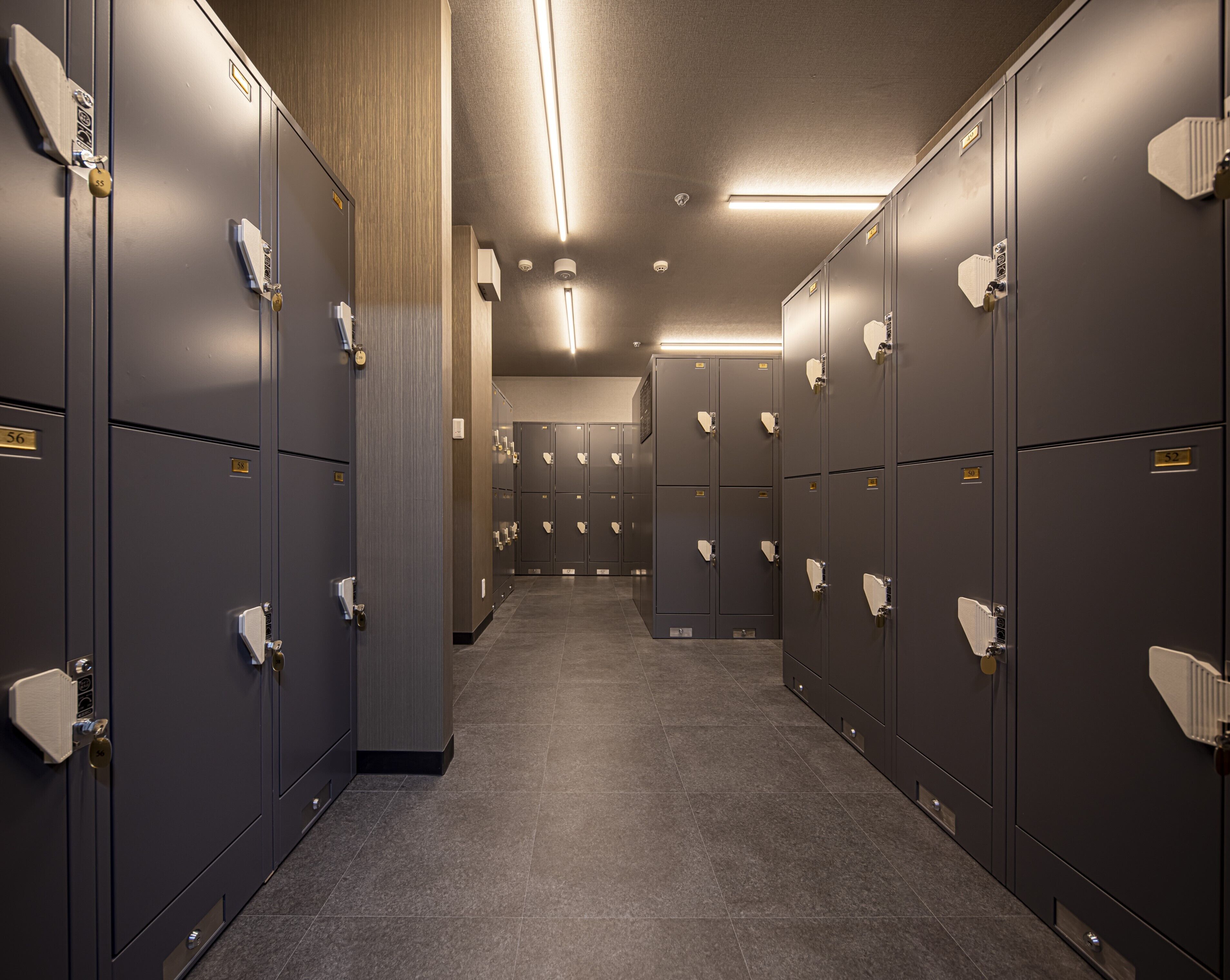 A row of lockers with numbers on the doors, a tiled floor, and a ceiling with linear lights.