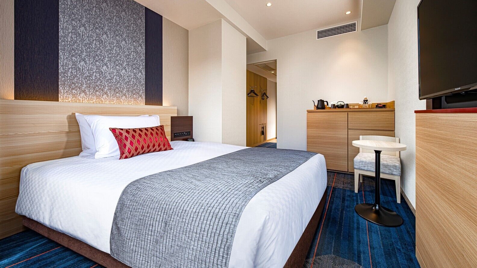 A hotel room with a large bed, a bedside table, a TV, a small table with chairs, and a wardrobe.