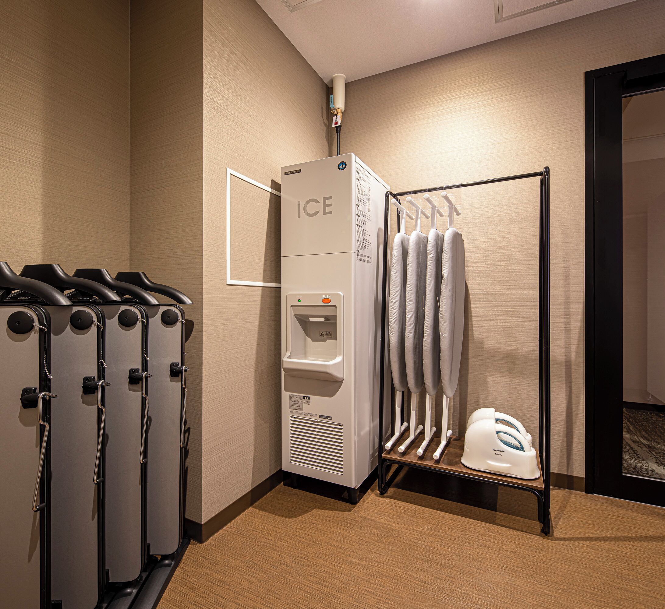 A hotel room with a wall-mounted ice dispenser, a rack of hanging towels, and a set of ironing boards.