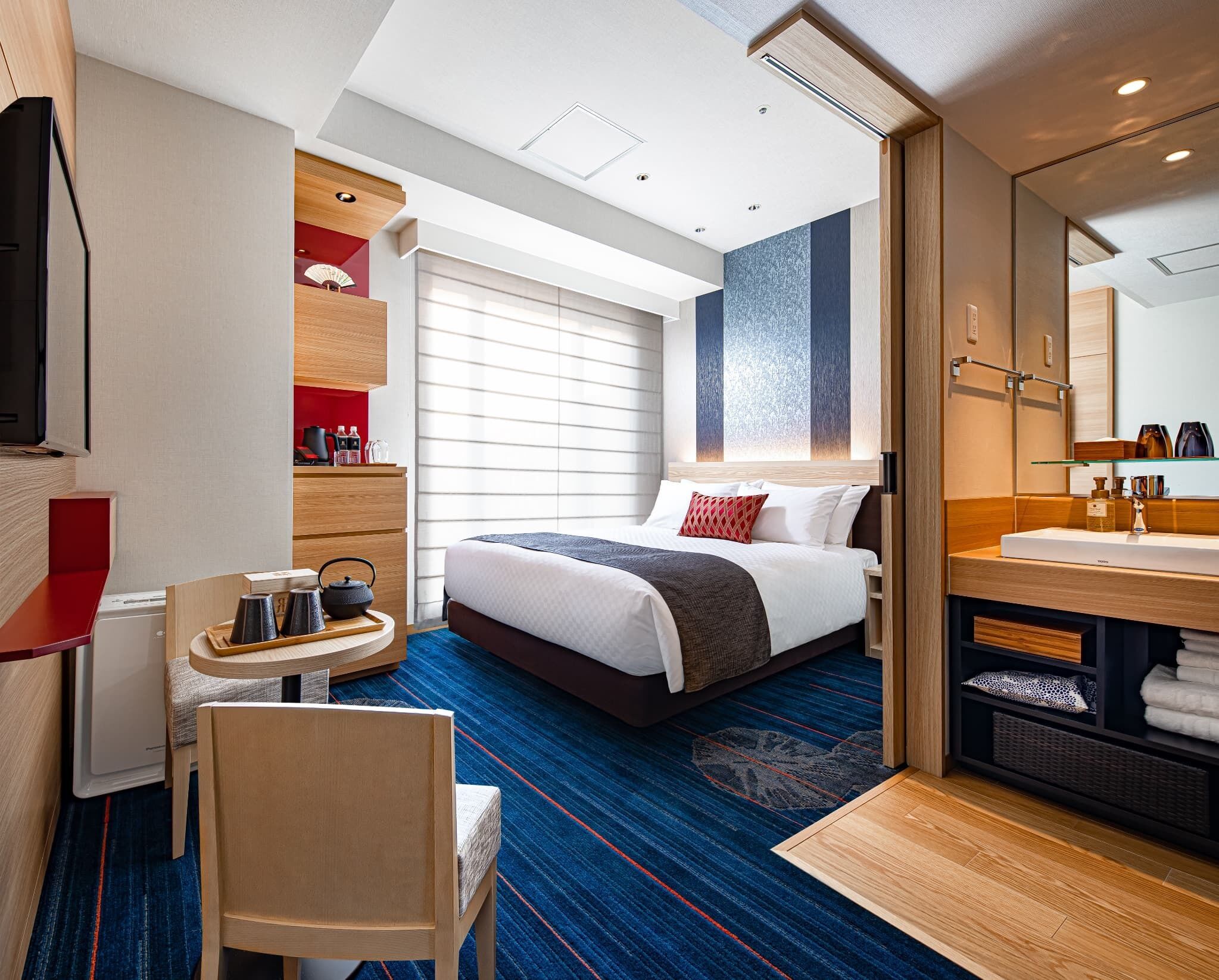 A hotel room with a bed, a desk with a chair, a bathroom with a mirror, and a window with blinds.