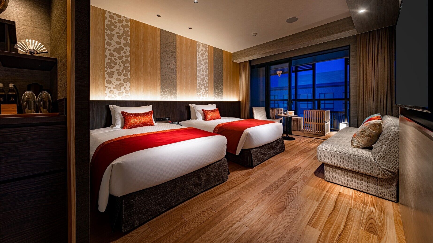 A hotel room with two beds, a sofa, a small table, and a view of the city at night.