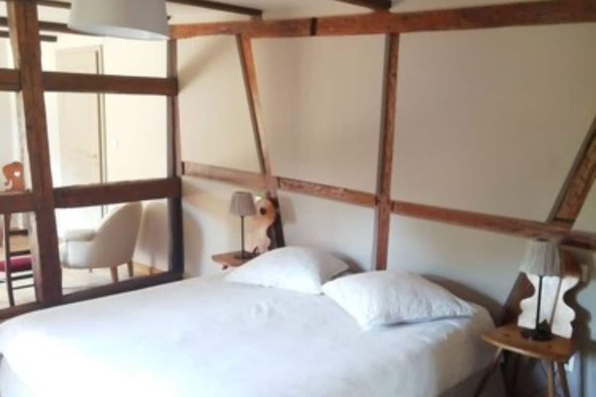 A bedroom with a large bed, wooden beams, a small table with a lamp, and a chair.