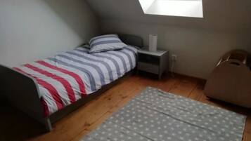 Une petite chambre avec un lit simple, un plafond en pente, une table de chevet, une lampe, un tapis orné de motifs en forme d’étoiles et une fenêtre donnant sur un immeuble.
