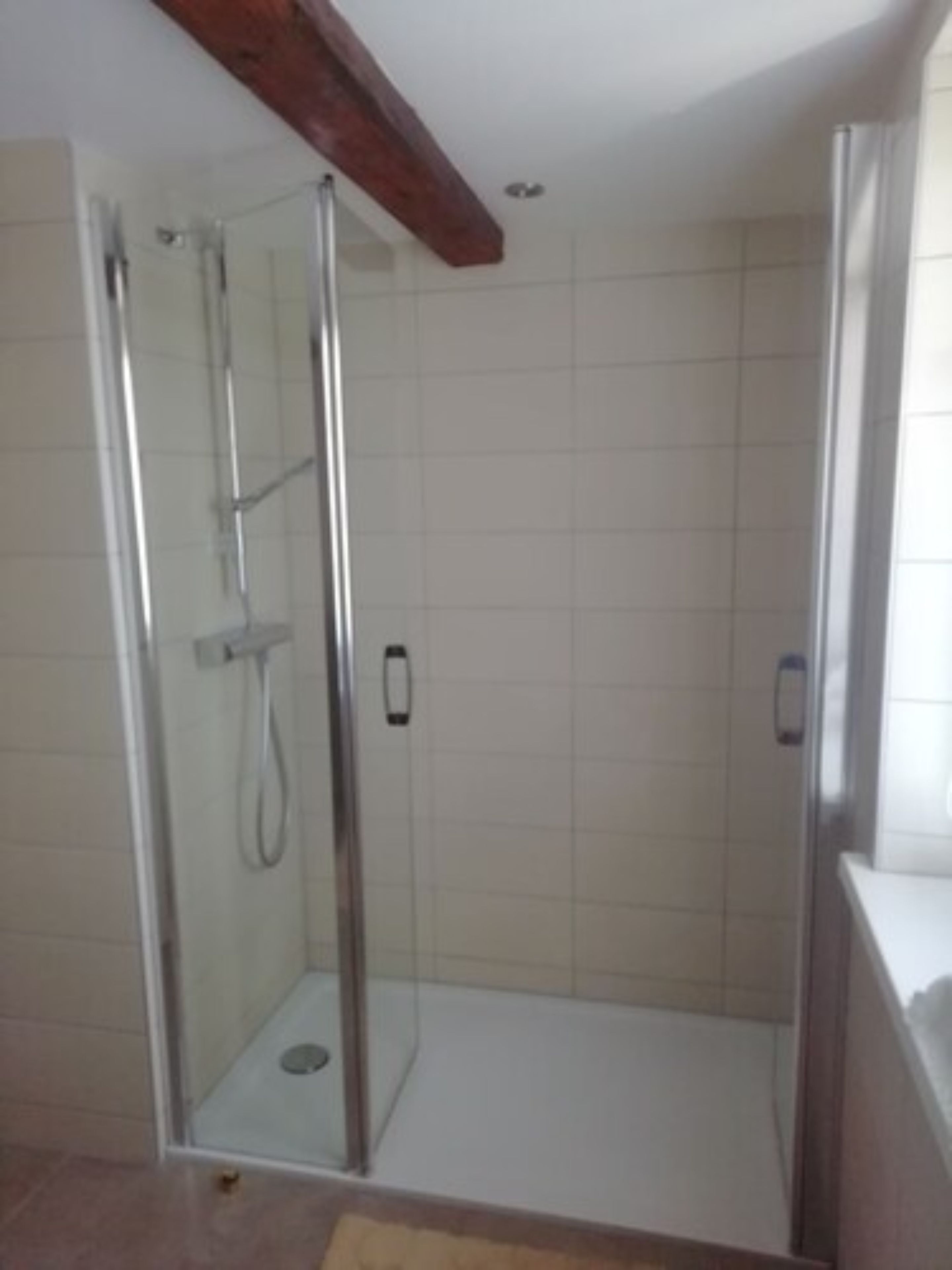 Ein Duschbereich mit Glaswänden, gefliestem Boden und einem Holzbalken an der Decke.