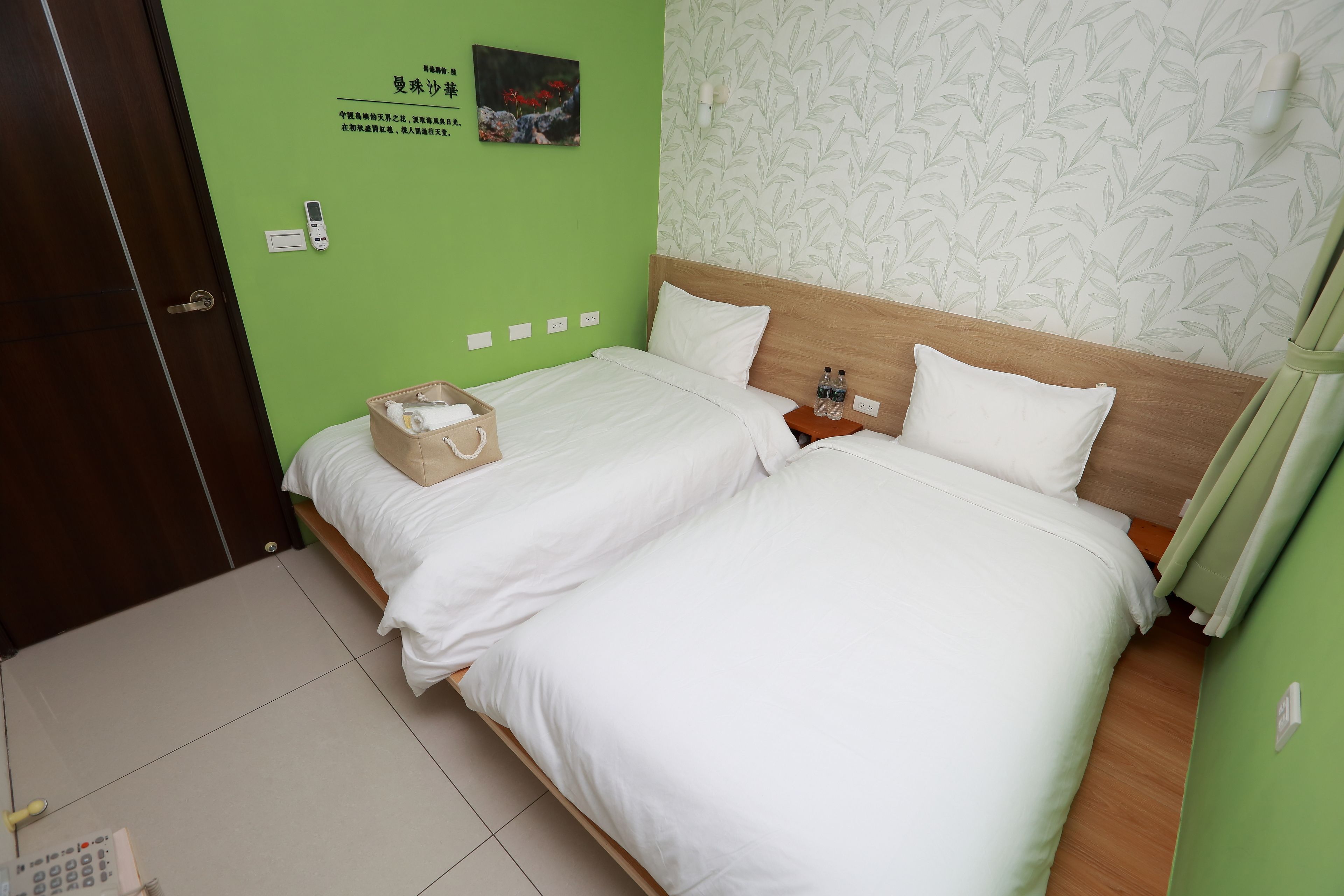 A hotel room with two beds, a wooden headboard, a green wall, a floral wallpaper, and a small bedside table with bottles.