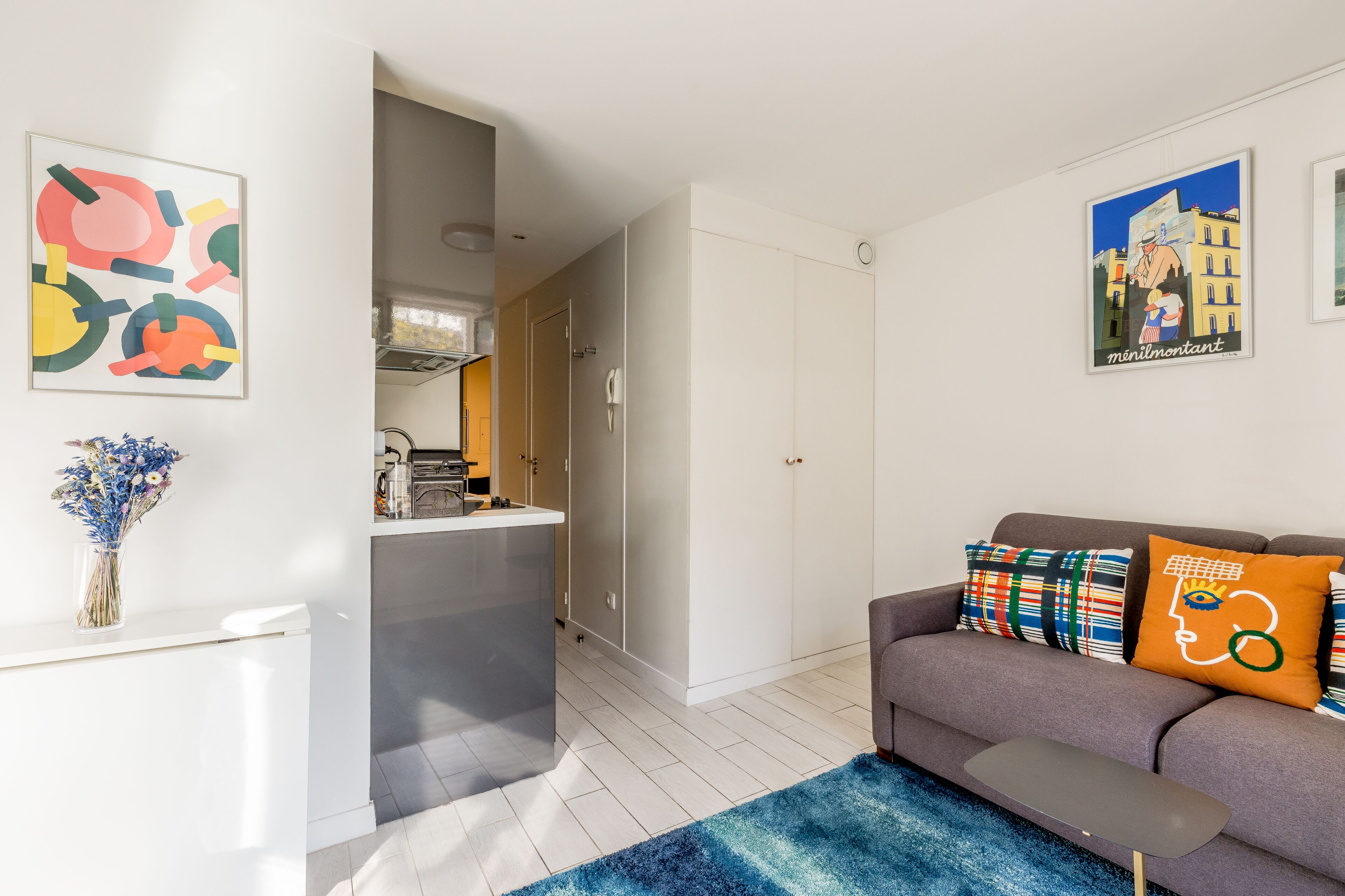 Een moderne woonkamer met een grijze bank, kleurrijke kussens, een blauw vloerkleed en kunstwerken aan de wanden.