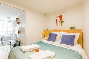 A neatly arranged hotel room with a bed, pillows, a bedside table with a lamp, a wall-mounted artwork, and a window with a view. - Résidence Louvre - Bellan (Paris)