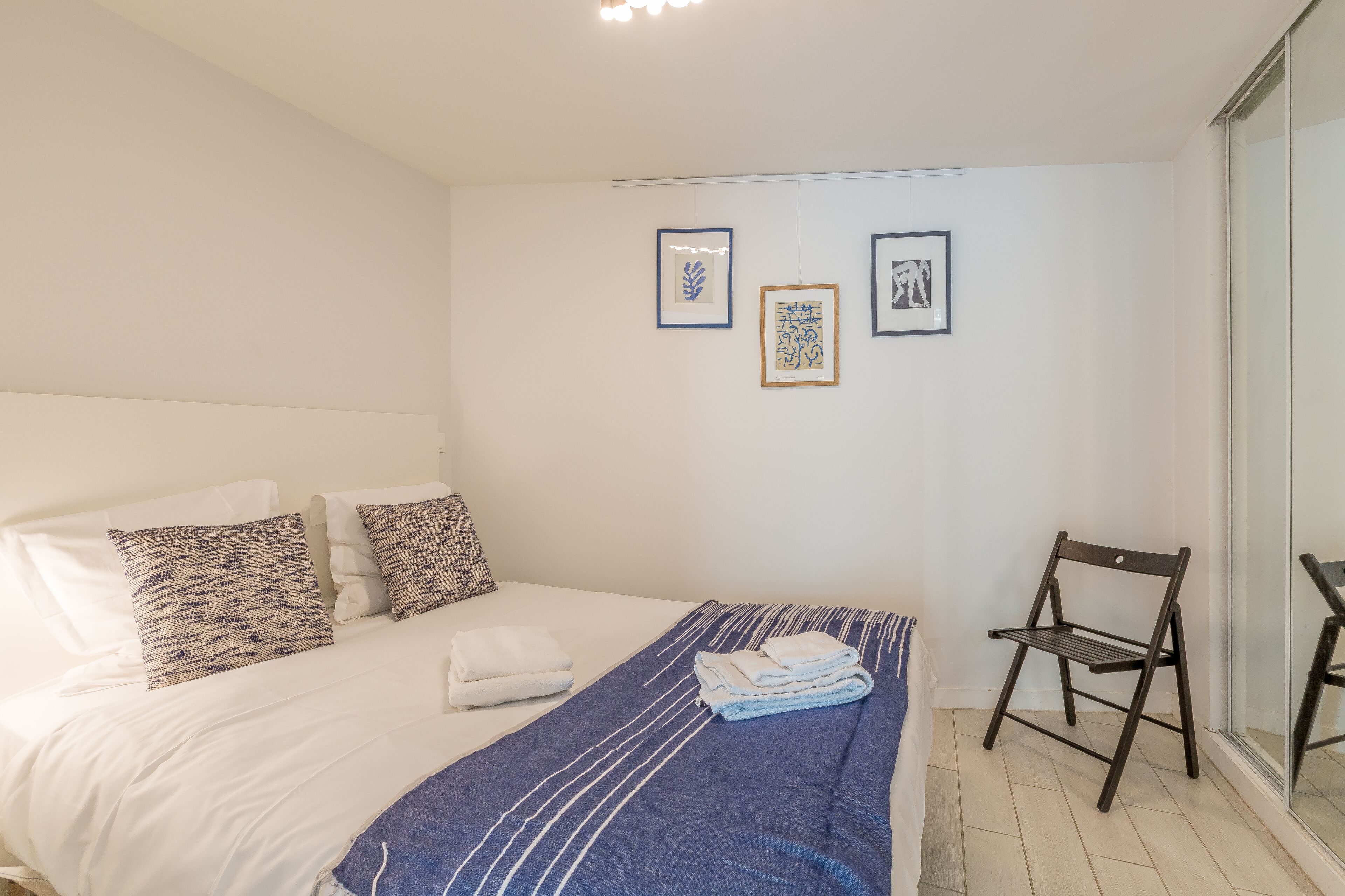 Een slaapkamer met een bed, kussens, een deken, een stoel en kunst aan de muur.
