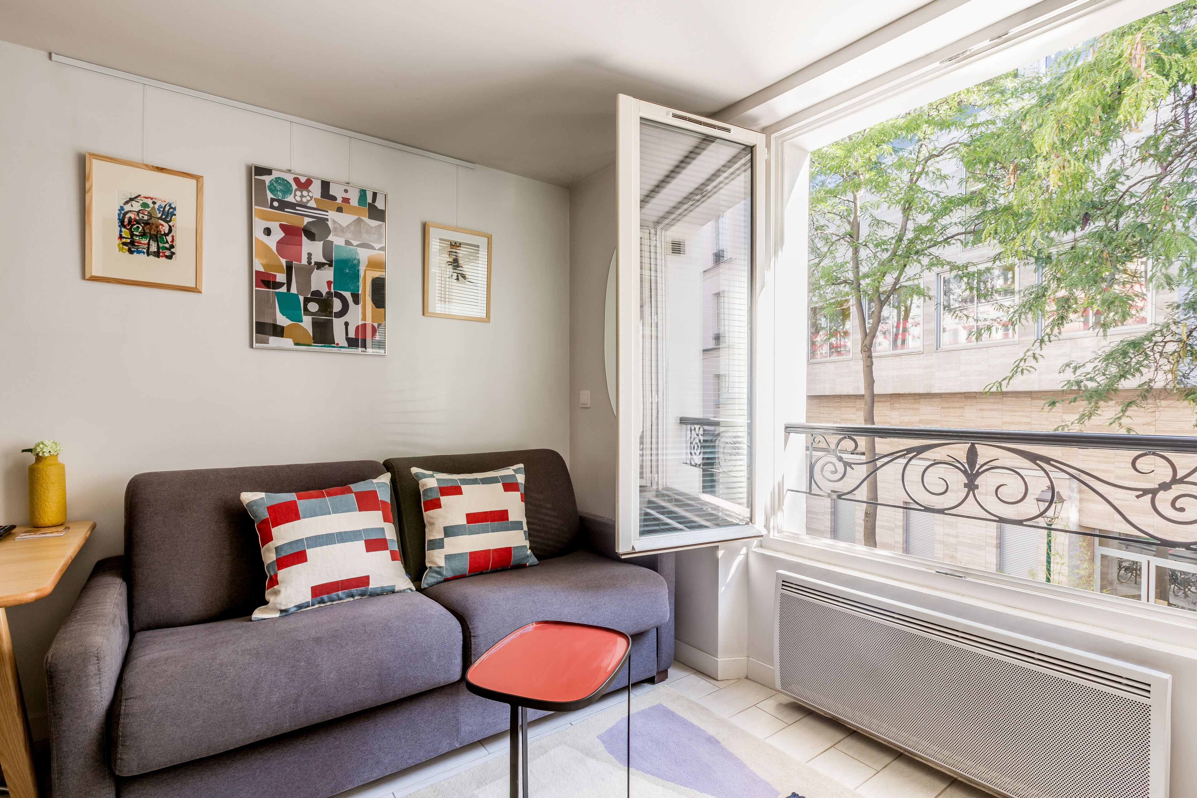 A modern living room with a grey sofa, two colorful pillows, a small red table, and a large window with a view of trees and buildings.