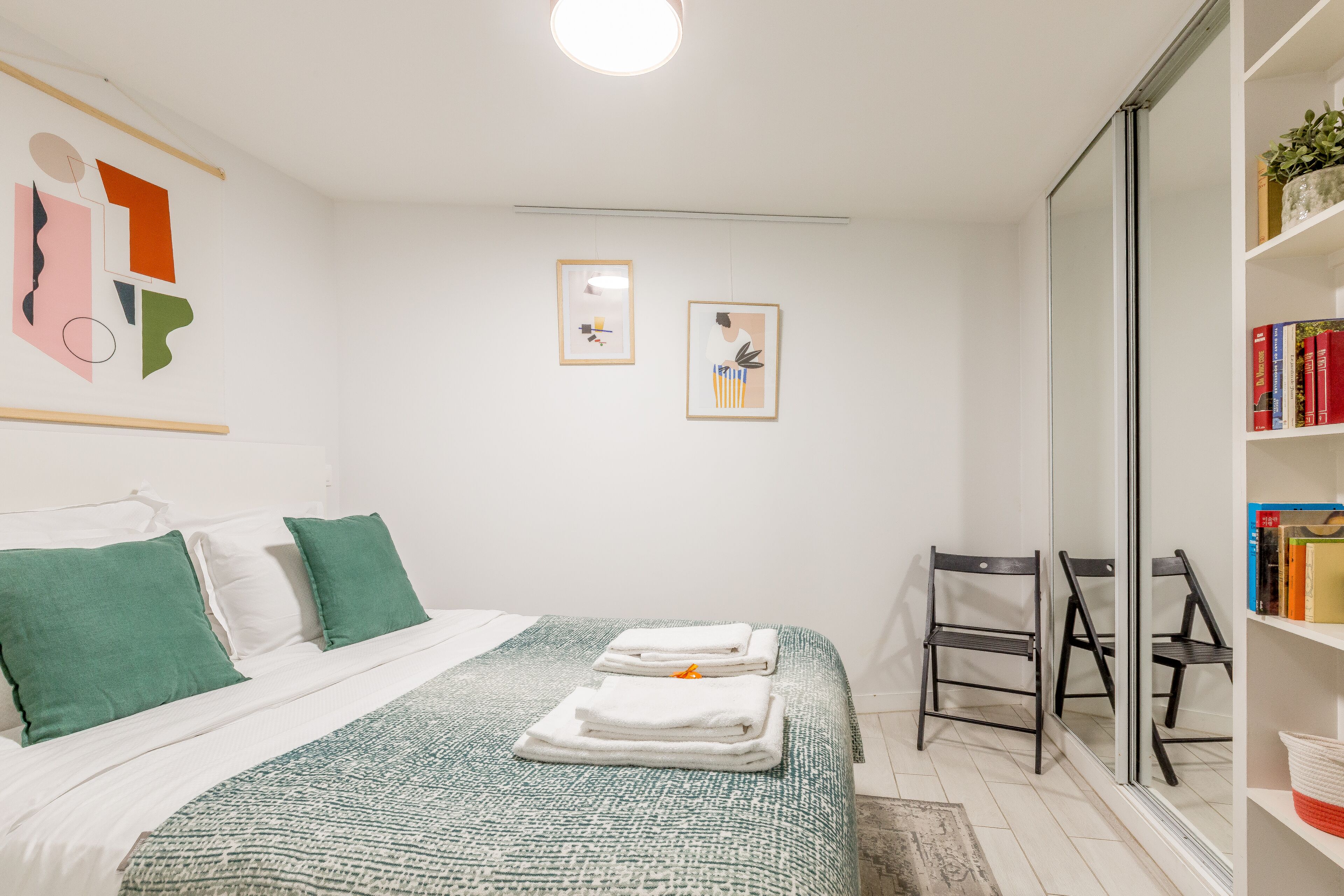 Ein Hotelzimmer mit einem Bett, grünen Kissen, einer weißen Decke, zwei gerahmten Bildern an der Wand, einem Bücherregal mit Büchern und einem Sessel.