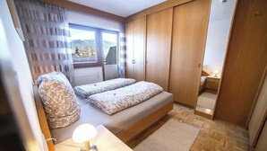 A bedroom with two beds, a nightstand, a lamp, a window with curtains, and a wardrobe. - Canisiusbrünnl (Rum)