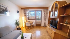 A cozy living room with a sofa, a dining table, and a TV. There is a window with curtains, a plant, and a clock on the wall. - Canisiusbrünnl (Rum)
