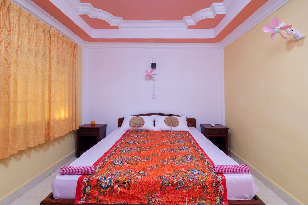 A bedroom with a large bed, two bedside tables, a ceiling fan, and a window with curtains.