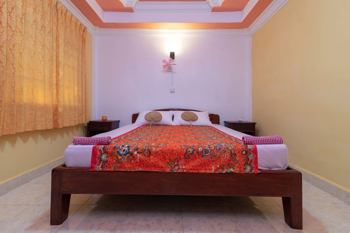A bedroom with a wooden bed, a patterned bedspread, and a wall-mounted fan.