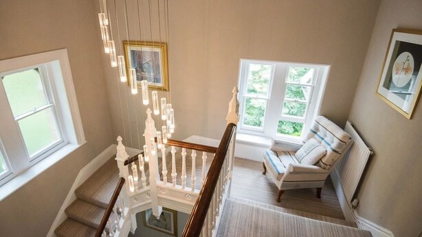 A staircase with a wooden handrail and white balusters, leading to a room with a beige wall, a window, and a chair with a cushion. - The Elmfield (Ilfracombe)