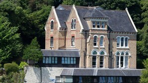 A large, multi-story building with a prominent tower and arched windows, surrounded by dense greenery. - The Elmfield (Ilfracombe)