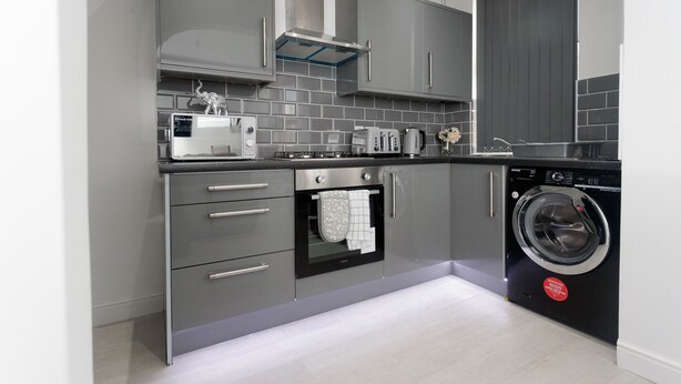 A modern kitchen with grey cabinets, a built-in oven, a washing machine, and a tiled backsplash. - Burton House (Leeds)
