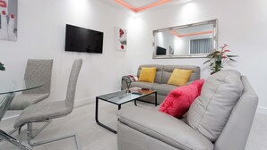 A modern living room with a glass coffee table, a grey sofa with a red pillow, a grey armchair, a flat-screen TV, and a large mirror. - Burton House (Leeds)