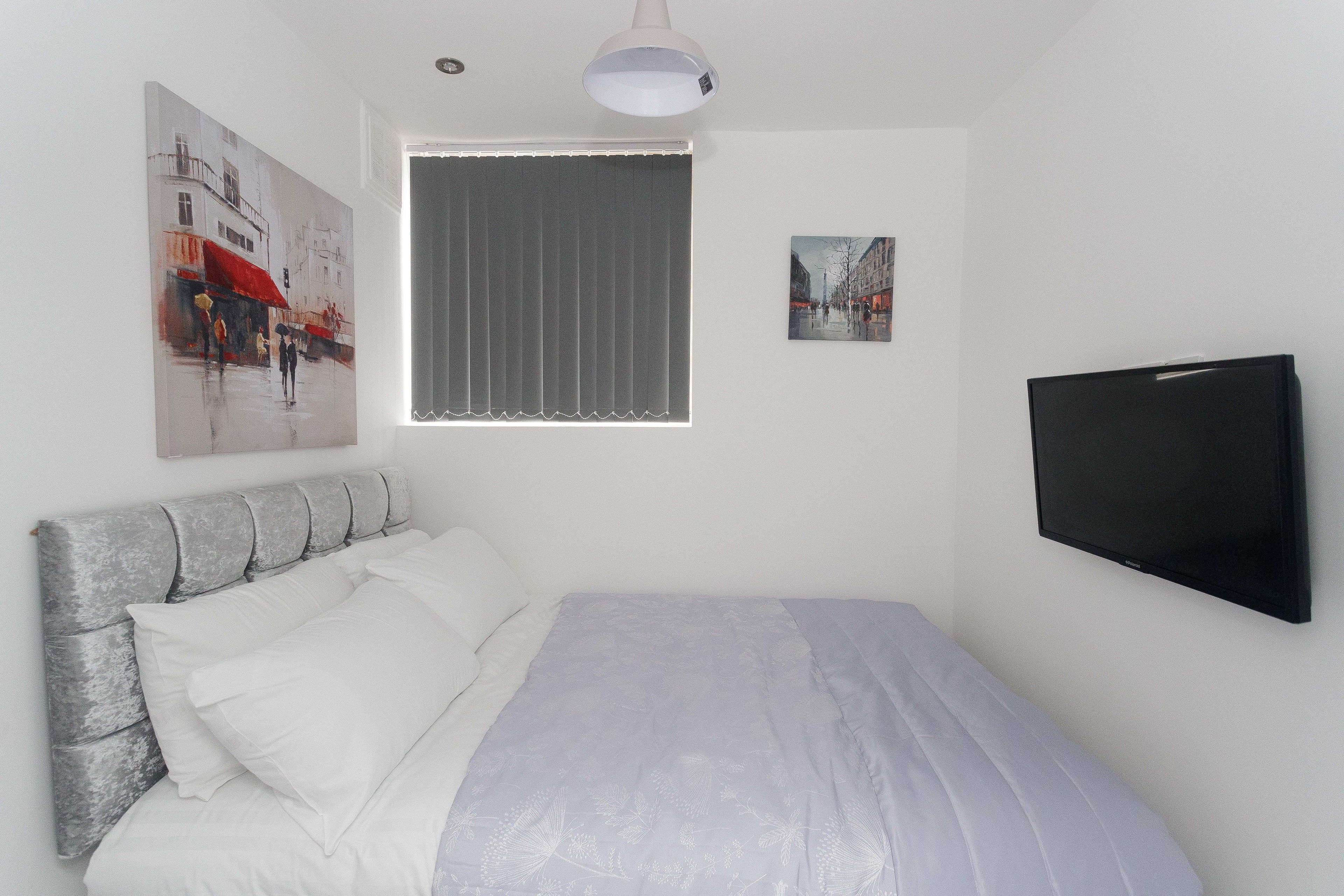 A bedroom with a bed, a window with blinds, a wall-mounted TV, and two paintings.