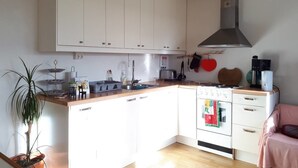 Eine kompakte Küche mit weißen Schränken, einem Herd, einer Spüle und einem kleinen Backofen. Links steht eine Pflanze und an der Wand hängt eine rote Herz-Dekoration.