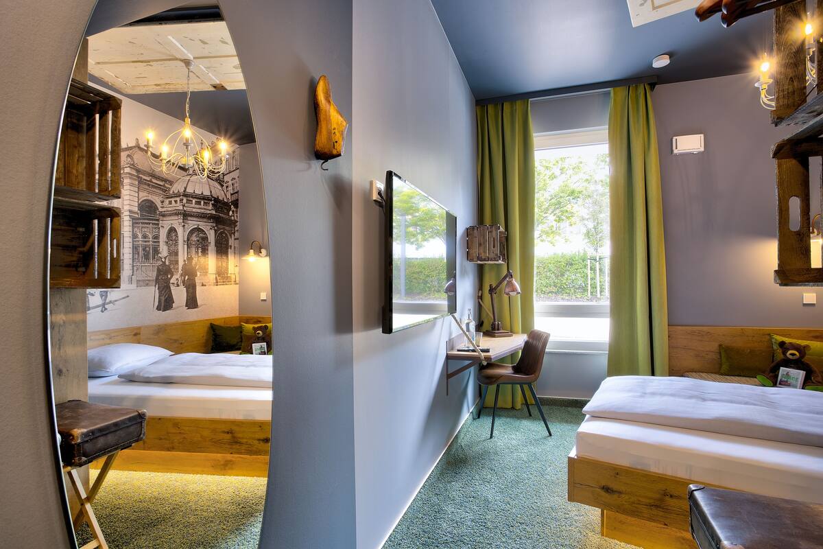 Habitación de hotel con dos camas, un escritorio, una silla, un ventanal con cortinas, un candelabro y un televisor montado en la pared.