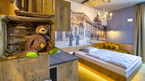 Una habitación de hotel con una cama grande, cabecera de madera, un osito de peluche y un mural de un edificio histórico.