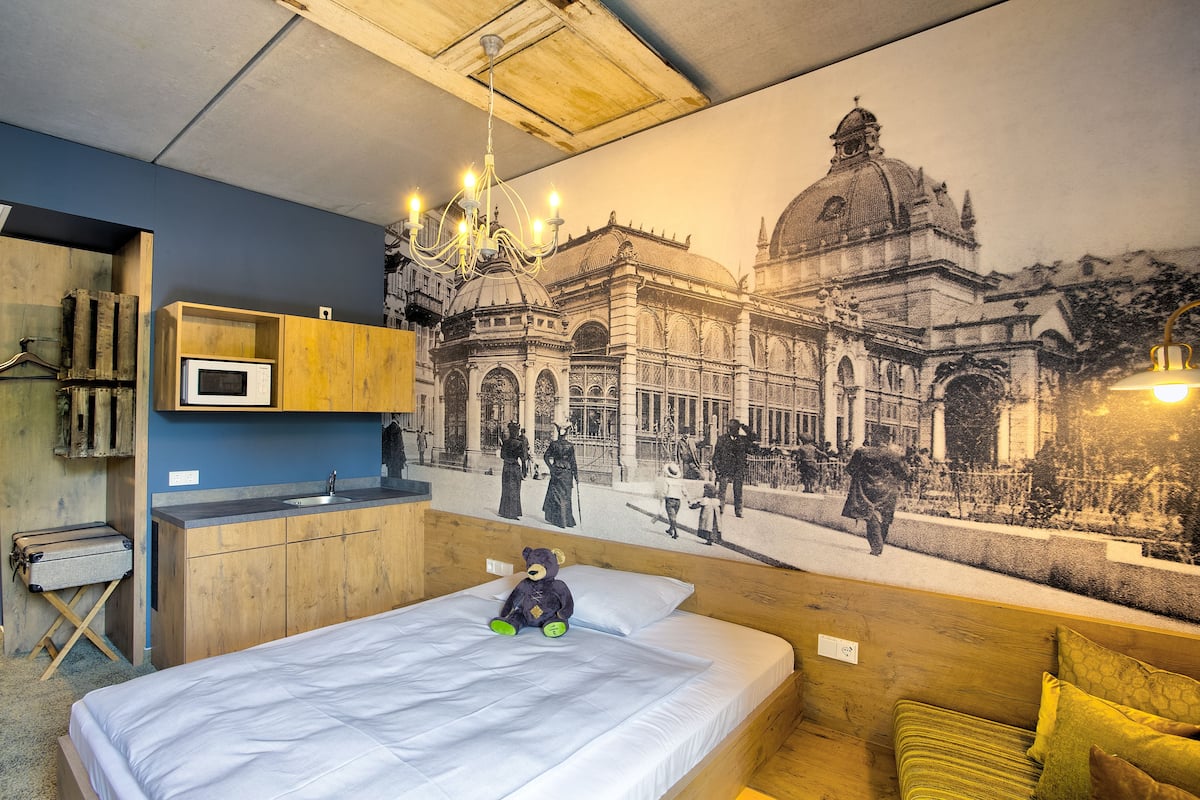 Una habitación de hotel con cama, cabecera de madera, un pequeño escritorio, una silla, una ventana y un mural en la pared.
