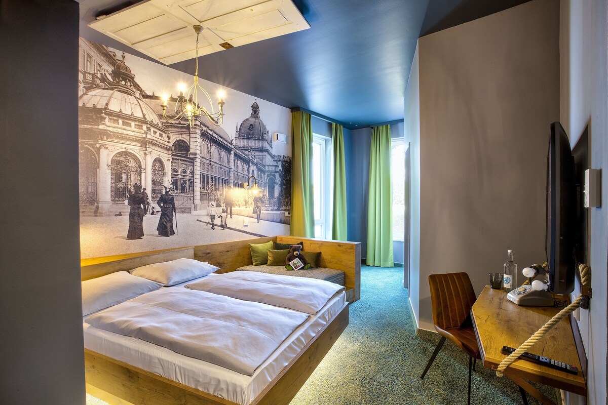 A hotel room with a large mural, a wooden bed, a desk with a chair, and a television.