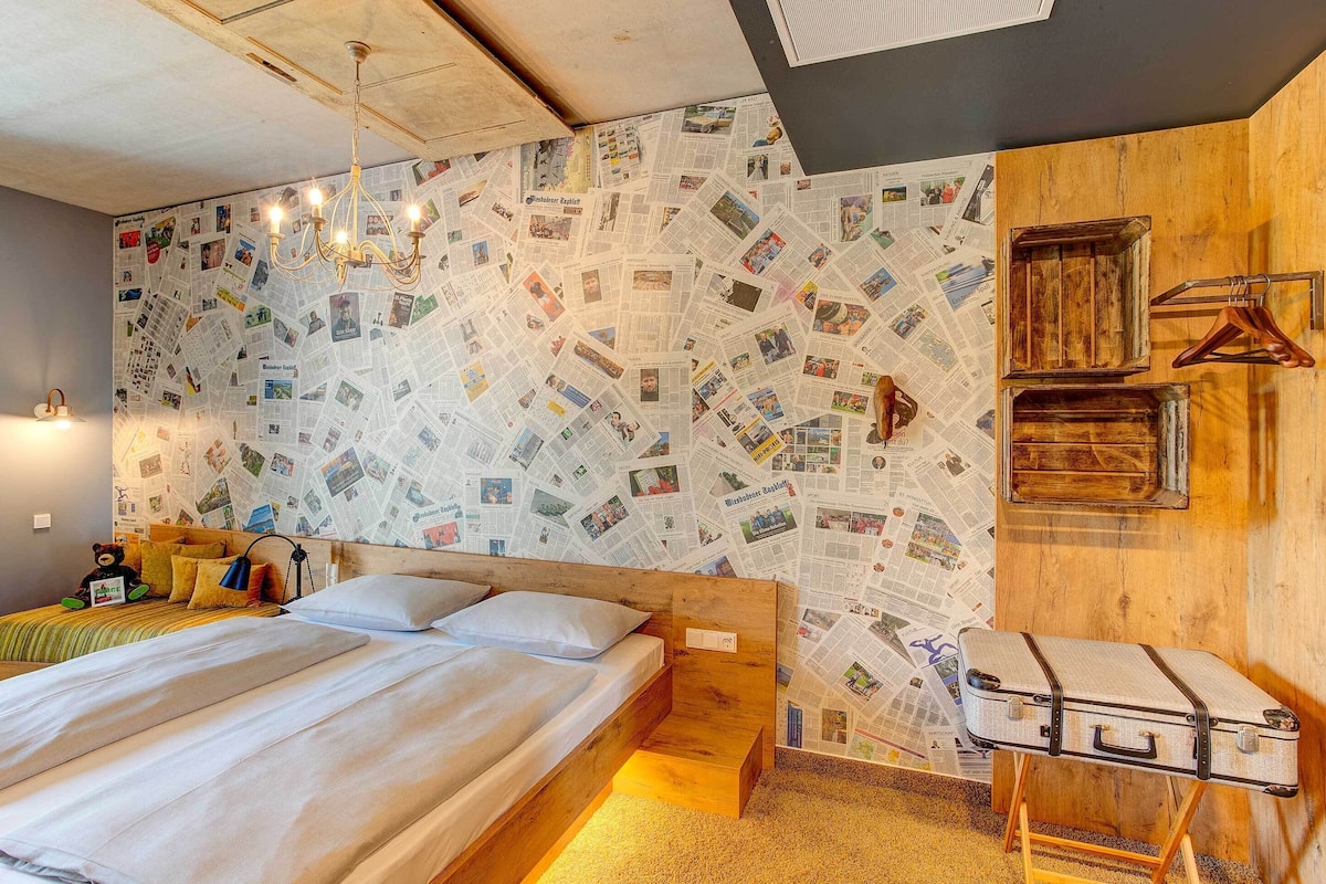 Un dormitorio con una cama, cabecera de madera, pared cubierta de recortes de periódicos, lámpara colgante y una maleta sobre un atril.