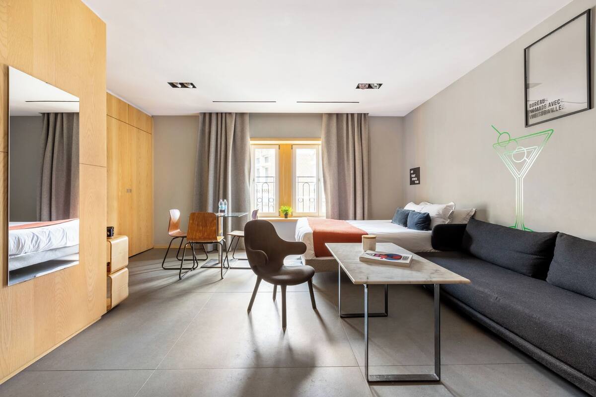 A modern hotel room with a bed, a sofa, a dining table, and a chair. There is a window with curtains, a wall-mounted TV, and a neon sign.
