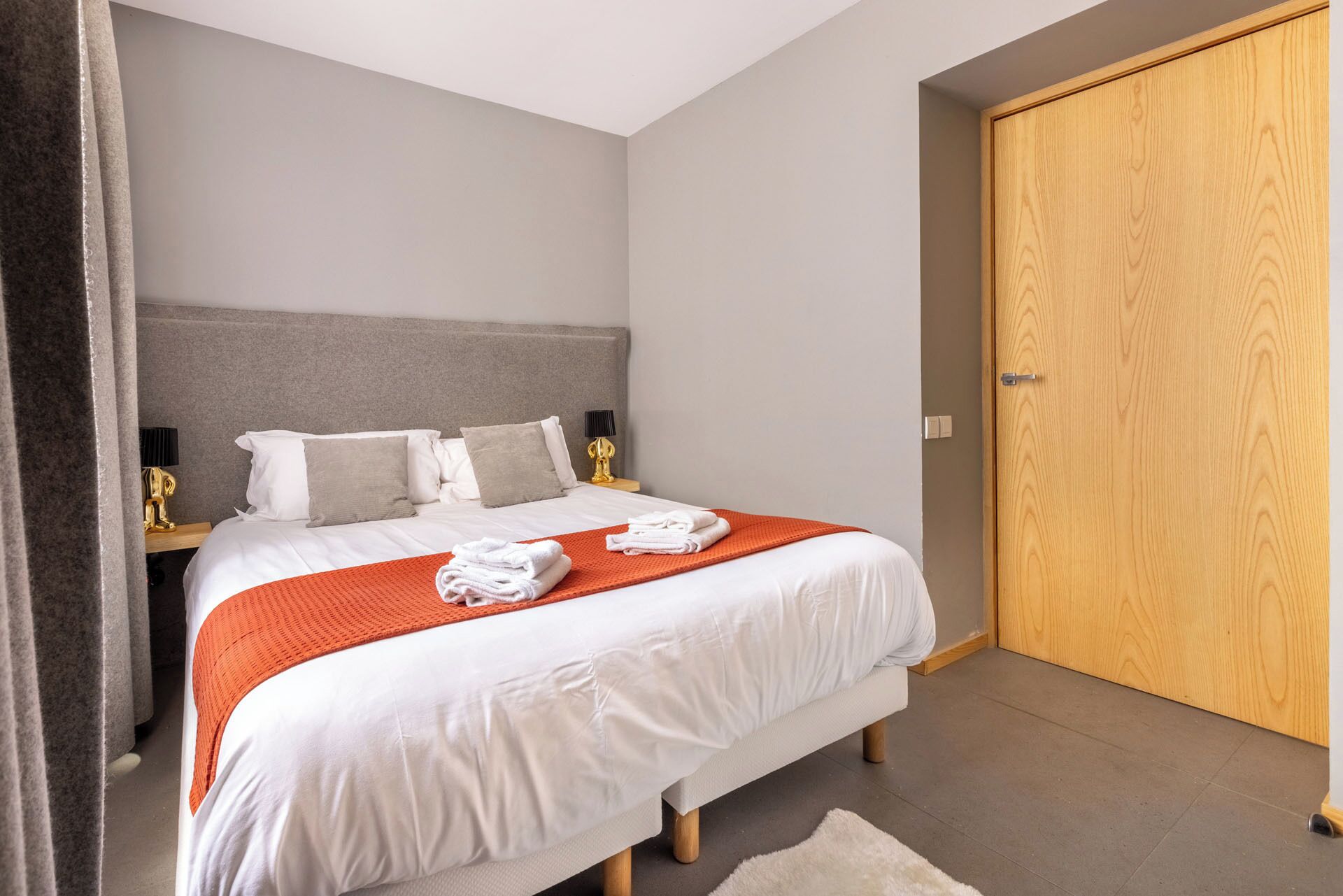 A neatly made bed with white linens and an orange throw blanket, a grey headboard, and a wooden wardrobe.