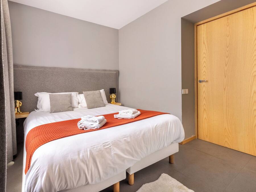 A neatly made bed with white linens and an orange throw blanket, a grey headboard, and a wooden wardrobe.
