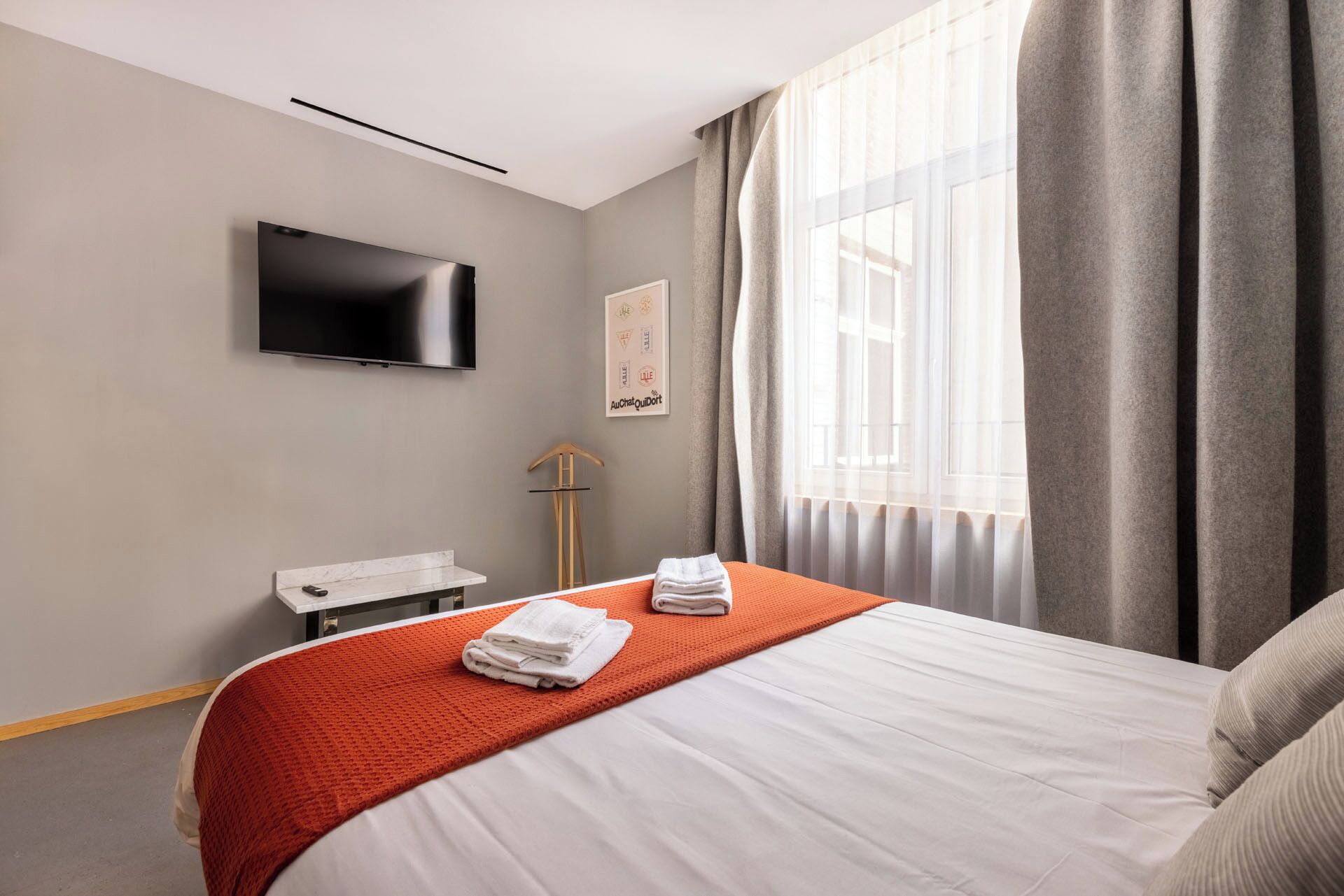 A hotel room with a bed, a TV mounted on the wall, a desk with a chair, and a window with curtains.