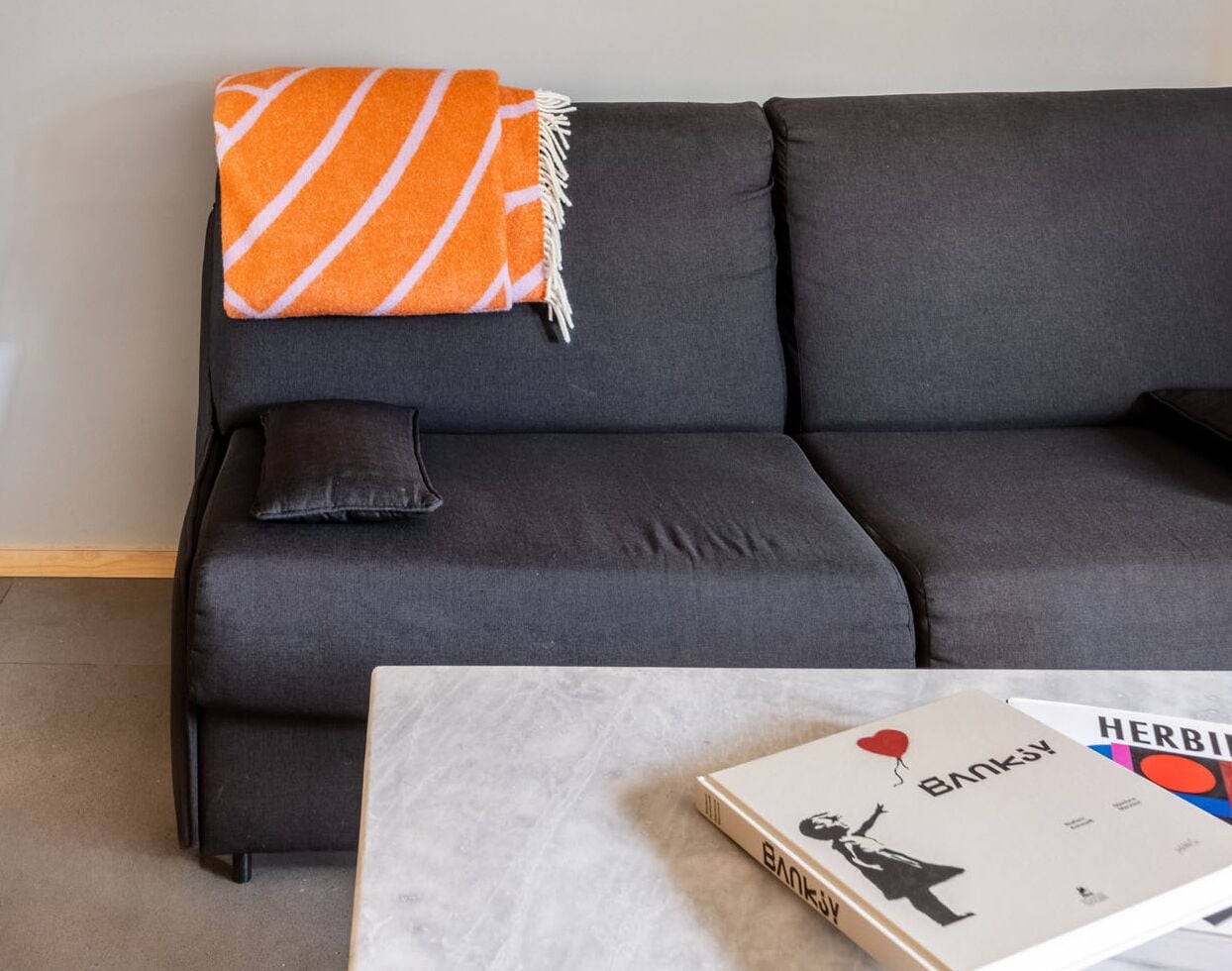 Un sofá gris oscuro con una manta anaranjada a rayas y una sola almohada negra. Delante del sofá, una mesa de centro blanca con un libro titulado “Banky” y un objeto pequeño negro.