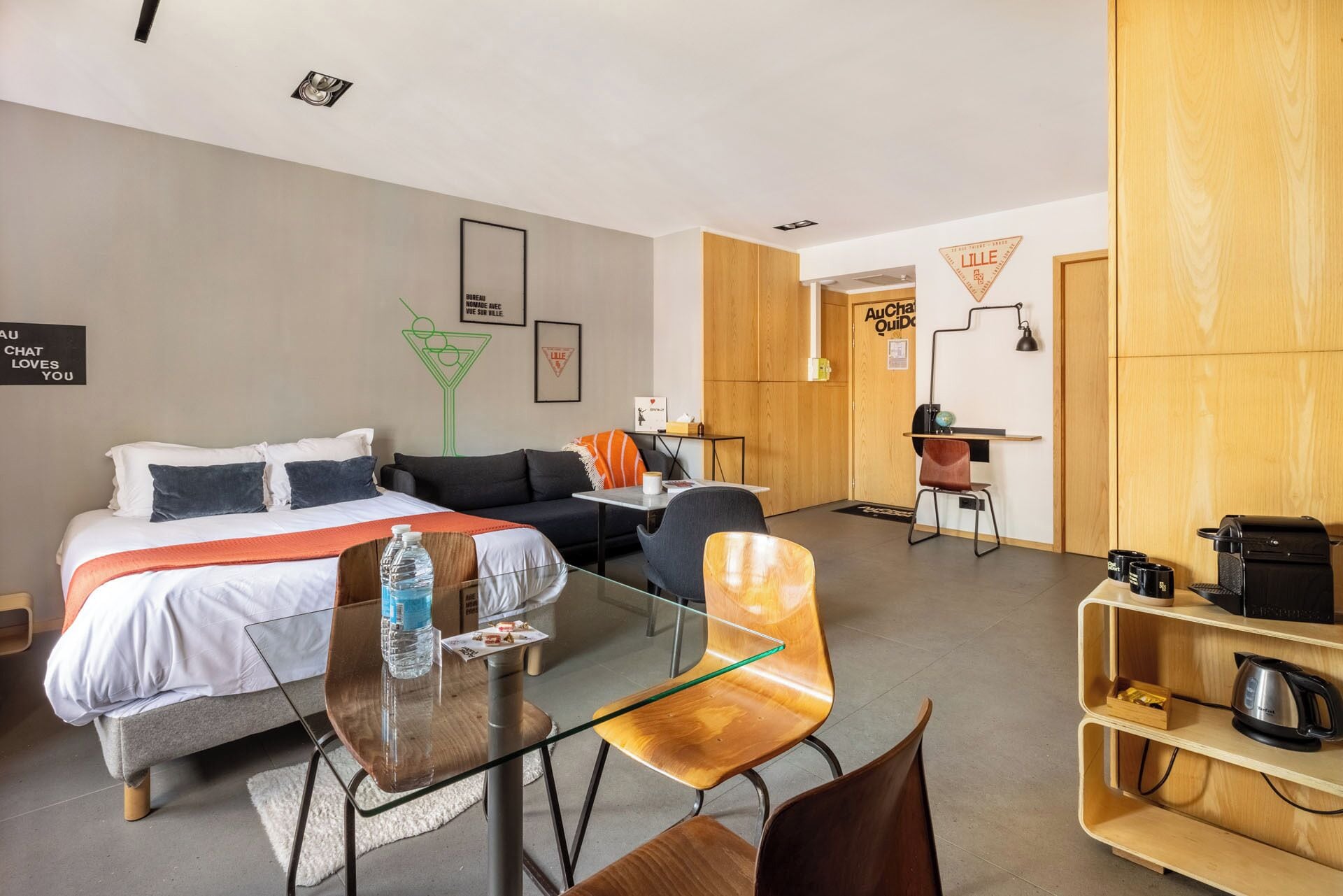Una camera d'albergo moderna con un letto, un tavolo da pranzo in vetro, una poltrona e un angolo cottura.