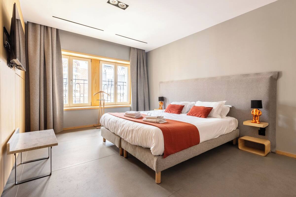 A modern hotel room with a large bed, a bedside table, a lamp, and a view of the city through the window.