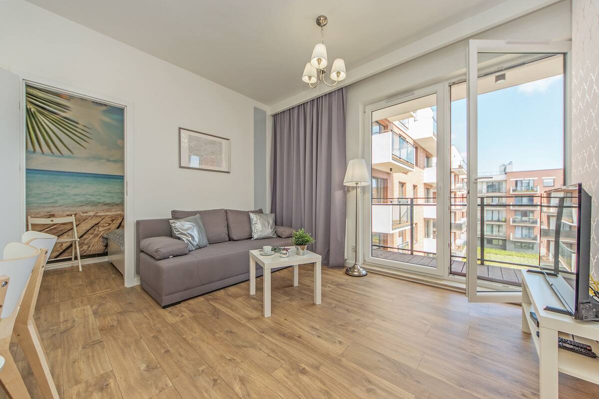 Un salon moderne avec un canapé gris, une petite table blanche et une grande fenêtre donnant sur des immeubles.