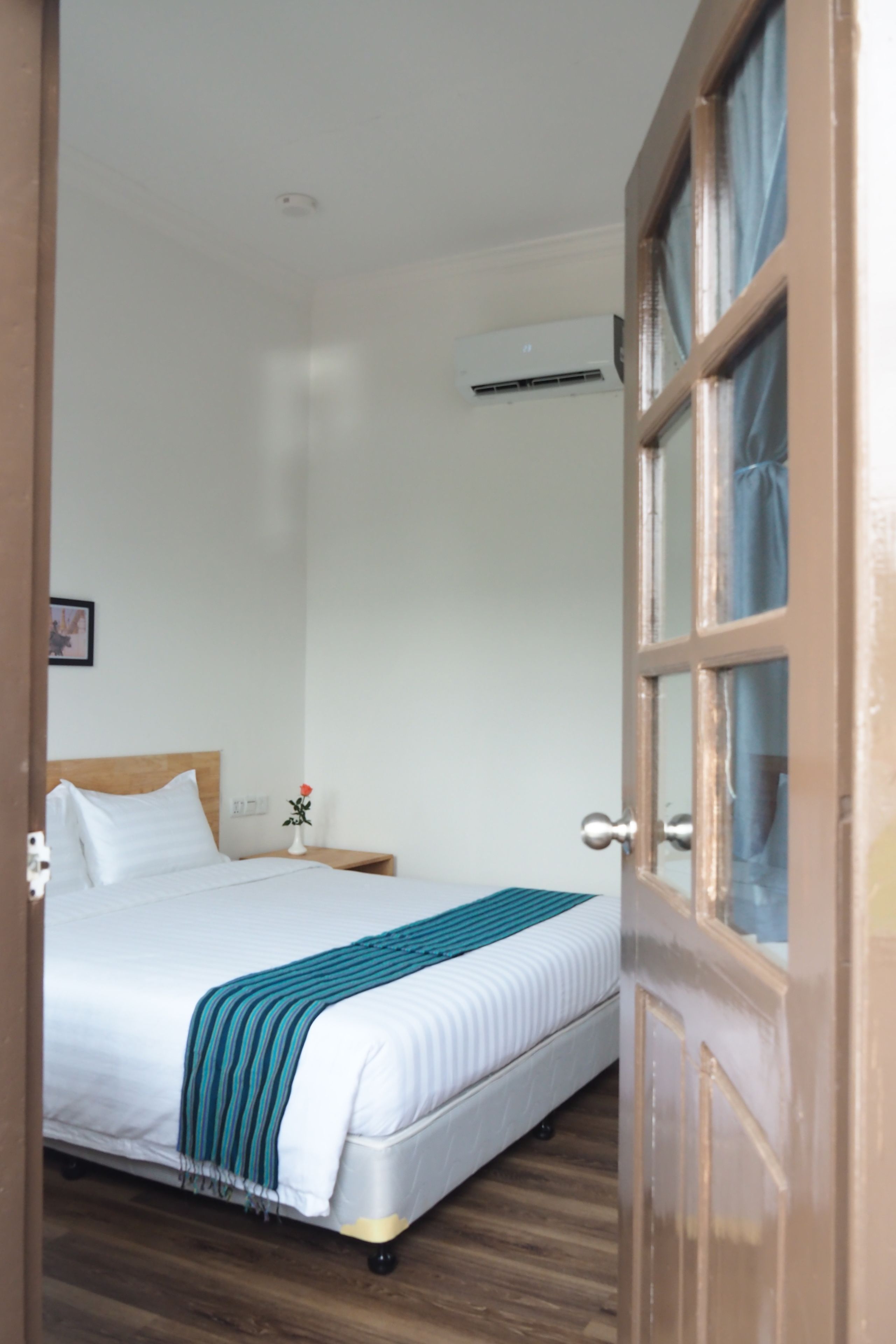 Een slaapkamer met een bed, houten vloer, nachtkastje, vaas met een bloem en een airconditioning.