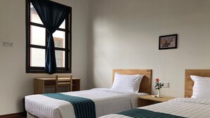 Habitación de hotel con dos camas, un escritorio de madera, una silla, una ventana con cortinas, un cuadro en la pared y un florero con una rosa.