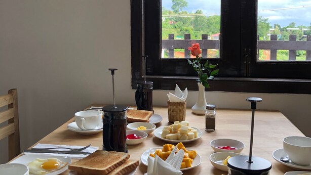 Una mesa de desayuno con diversos alimentos y una cafetera francesa, frente a una ventana con vista a follaje.