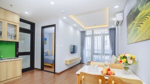 Una habitación de hotel moderna con una mesa de comedor de madera, sillas, una pequeña cocina, una cama y un gran ventanal con cortinas.
