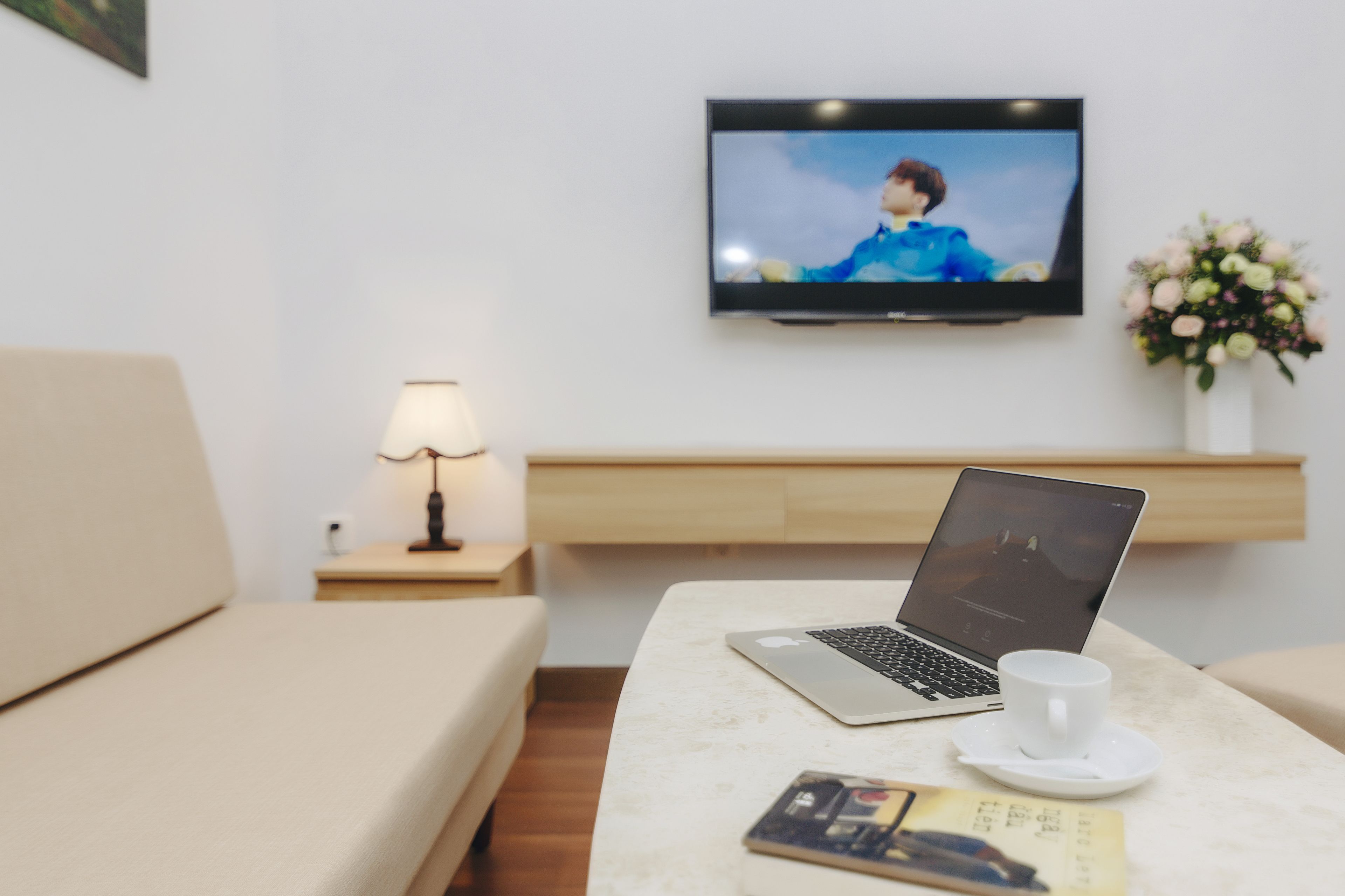 A hotel room with a bed, a desk with a laptop, a TV, a lamp, and a flower arrangement.