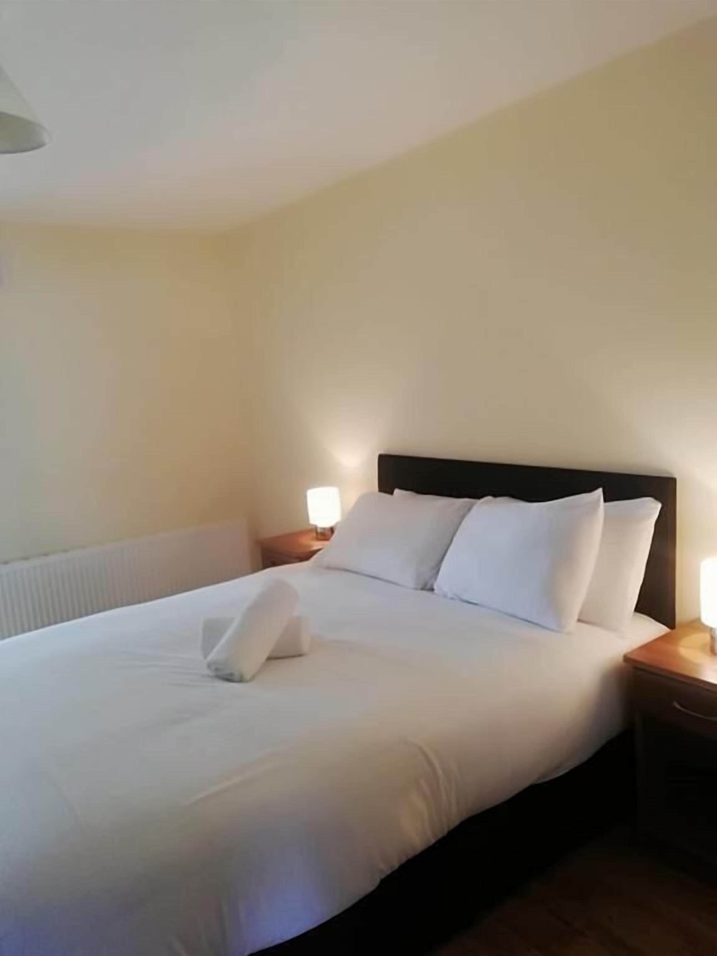 A neatly made bed with white linens and pillows, a wooden headboard, and bedside lamps on wooden nightstands.