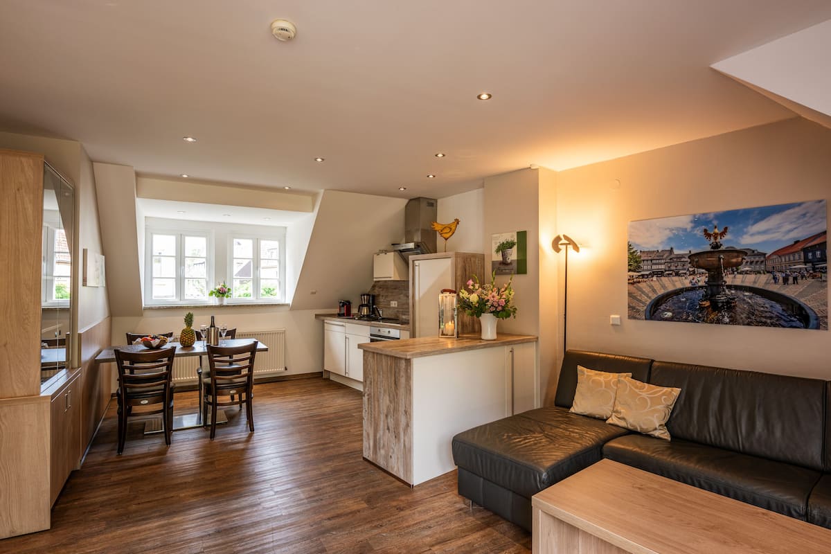 Een moderne keuken met houten kastjes, een eethoek met een tafel en stoelen, een zwarte leren bank en een schilderij van een Europees stadsplein.