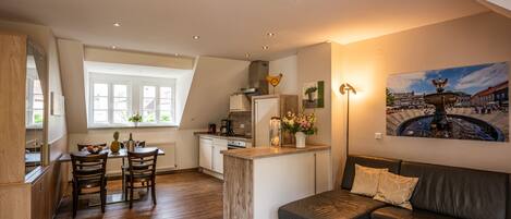 A modern kitchen with wooden cabinets, a dining area with a table and chairs, a black leather sofa, and a painting of a European town square.