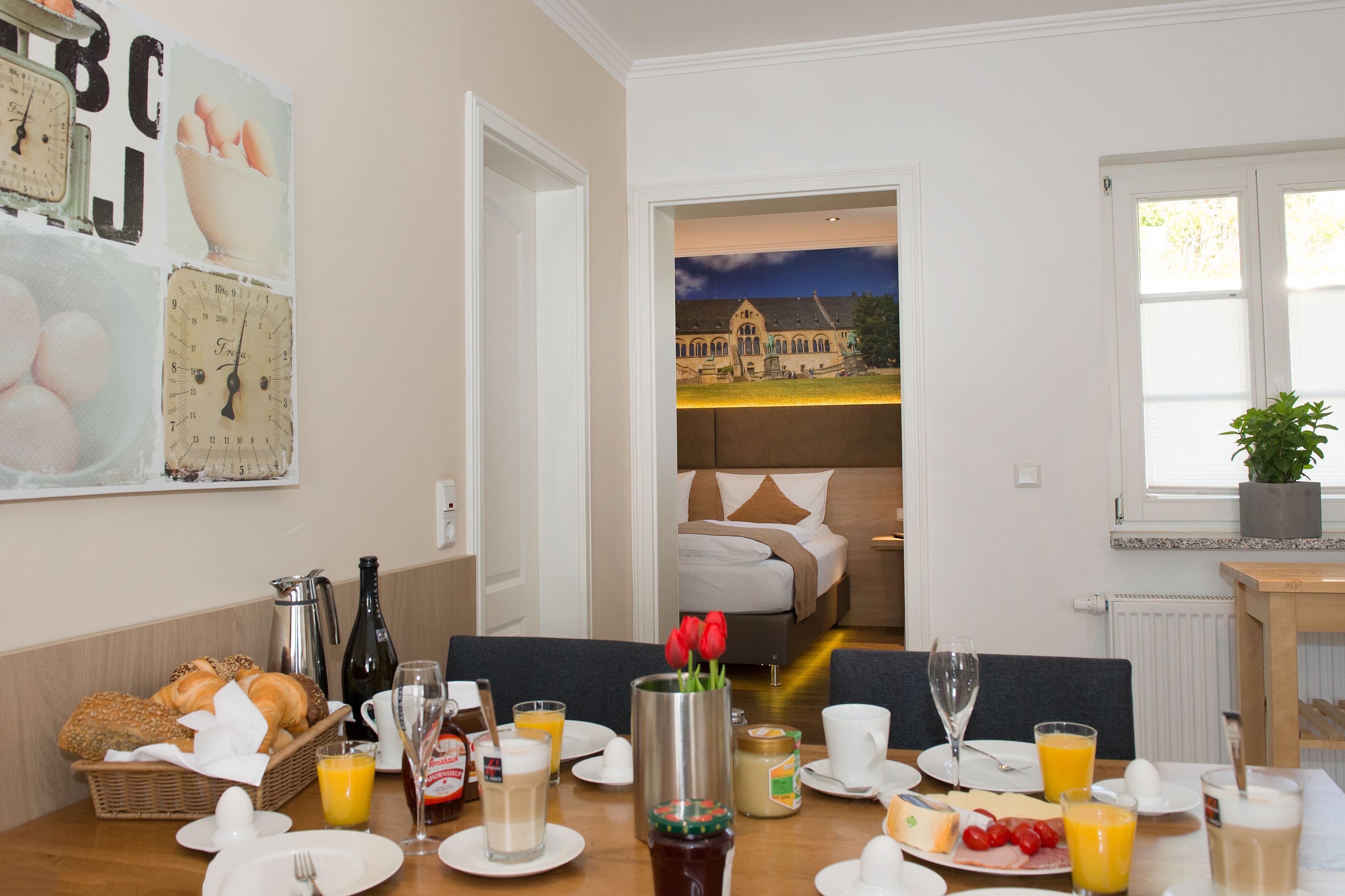 A breakfast table set with various food items and drinks, a vase with flowers, and a view of a bedroom through an open door.