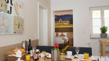 A breakfast table set with various food items and drinks, a vase with flowers, and a view of a bedroom through an open door.